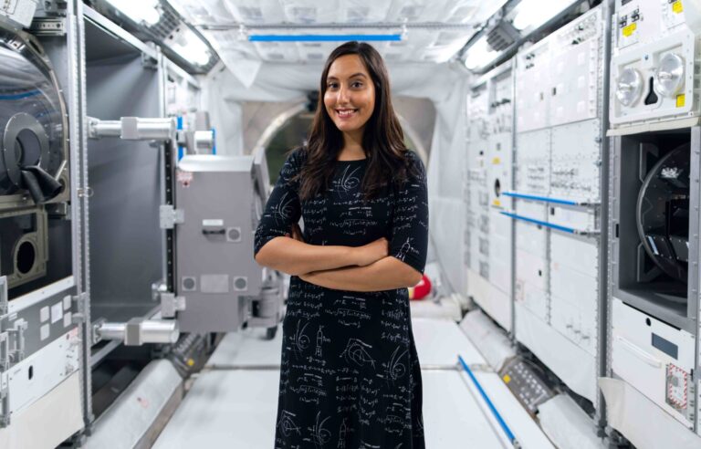 Mujer científica posa dentro de un laboratorio aeroespacial rodeada de equipos tecnológicos y sistemas de investigación avanzada.